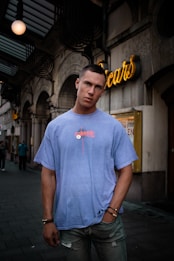 A young man stands in an urban street setting, wearing a blue T-shirt and jeans. Neon signage decorates the background, with architectural elements like arches and ornate details visible on the building. The man has a short haircut and is accessorized with bracelets and a necklace.