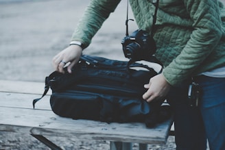 A person wearing a green quilted jacket is adjusting a black backpack on a wooden table. A camera hangs from the person's neck, indicating a possible photography outing or preparation.