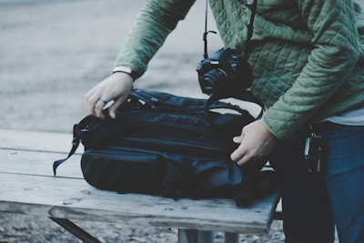 A person wearing a green quilted jacket is adjusting a black backpack on a wooden table. A camera hangs from the person's neck, indicating a possible photography outing or preparation.