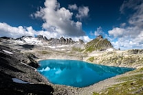 A stunning alpine landscape featuring a vivid blue lake surrounded by rocky, snow-capped mountains under a partly cloudy sky. The water reflects the surrounding peaks and sky, creating a serene atmosphere. Sparse patches of snow are visible on the mountainsides, and the terrain is rugged with scattered rocks and sparse greenery.