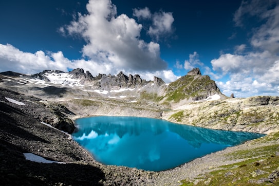 A stunning alpine landscape featuring a vivid blue lake surrounded by rocky, snow-capped mountains under a partly cloudy sky. The water reflects the surrounding peaks and sky, creating a serene atmosphere. Sparse patches of snow are visible on the mountainsides, and the terrain is rugged with scattered rocks and sparse greenery.