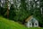 A small wooden cabin with a red roof is nestled among tall, dense evergreen trees. The cabin features a small balcony with a colorful flag and rustic wooden decorations. It sits on a lush green grassy slope surrounded by a wooden fence, evoking a serene and secluded atmosphere.