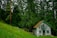 A small wooden cabin with a red roof is nestled among tall, dense evergreen trees. The cabin features a small balcony with a colorful flag and rustic wooden decorations. It sits on a lush green grassy slope surrounded by a wooden fence, evoking a serene and secluded atmosphere.