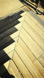 Wooden deck panels align to form a geometric pattern, casting sharp shadows under bright sunlight. A bicycle wheel partially visible at the edge adds an urban outdoor element.