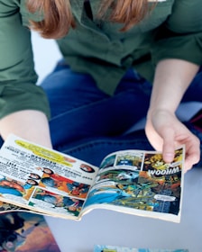 A person wearing a green shirt and blue jeans is holding an open comic book. The comic book features colorful illustrations and bold text, typical of graphic novels or superhero stories. The person's face is not visible, focusing attention on the vivid comic panels.