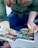 A person wearing a green shirt and blue jeans is holding an open comic book. The comic book features colorful illustrations and bold text, typical of graphic novels or superhero stories. The person's face is not visible, focusing attention on the vivid comic panels.