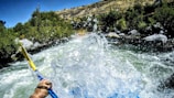 A scene of white water rafting with a kayak paddle in hand, splashing through a fast-flowing river surrounded by lush greenery and rocky banks under a clear blue sky.