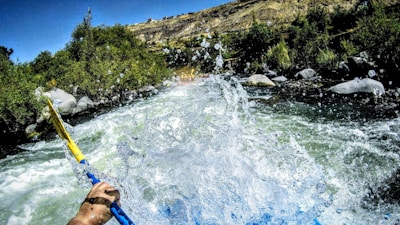 A scene of white water rafting with a kayak paddle in hand, splashing through a fast-flowing river surrounded by lush greenery and rocky banks under a clear blue sky.