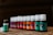 An array of small bottles with white caps are lined up in a diagonal row. Each bottle has a different colored label ranging from green, orange, red to purple. They appear to be essential oil bottles, with one prominently labeled 'Peppermint' in the foreground.