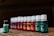 An array of small bottles with white caps are lined up in a diagonal row. Each bottle has a different colored label ranging from green, orange, red to purple. They appear to be essential oil bottles, with one prominently labeled 'Peppermint' in the foreground.