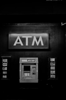A black and white photograph of an ATM machine embedded in a wall. Above the ATM, there is a prominent sign with large letters spelling 'ATM'. To the sides, there are advertisements or logos for various card networks.