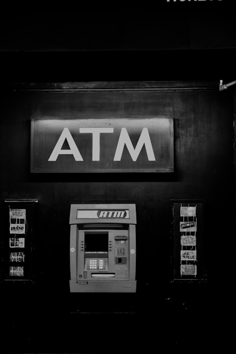 A black and white photograph of an ATM machine embedded in a wall. Above the ATM, there is a prominent sign with large letters spelling 'ATM'. To the sides, there are advertisements or logos for various card networks.