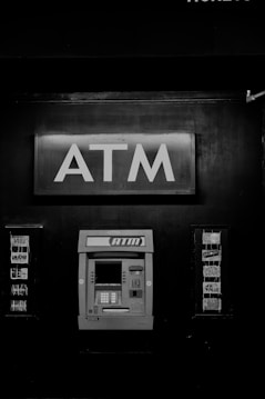 A black and white photograph of an ATM machine embedded in a wall. Above the ATM, there is a prominent sign with large letters spelling 'ATM'. To the sides, there are advertisements or logos for various card networks.