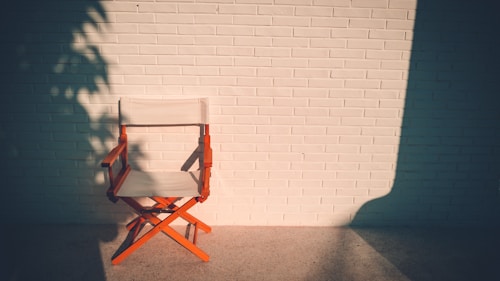 A wooden director's chair with a white canvas seat and backrest is placed against a white brick wall. Shadows of tree leaves and the chair are cast on the wall and ground, suggesting the lighting is from the side, likely from the setting sun.