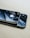 Two smartphones are placed on top of each other on a light wooden surface. The top phone features a prominent logo and a shiny black back, while the bottom phone displays a dual camera setup. Both devices have reflective surfaces, capturing glimpses of the surrounding environment.