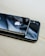 Two smartphones are placed on top of each other on a light wooden surface. The top phone features a prominent logo and a shiny black back, while the bottom phone displays a dual camera setup. Both devices have reflective surfaces, capturing glimpses of the surrounding environment.