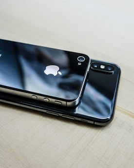 Two smartphones are placed on top of each other on a light wooden surface. The top phone features a prominent logo and a shiny black back, while the bottom phone displays a dual camera setup. Both devices have reflective surfaces, capturing glimpses of the surrounding environment.