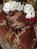 Hands adorned with intricate henna designs rest gently on a richly embroidered fabric. The wrists are decorated with bright floral bracelets, featuring white and red flowers, and the person is wearing golden rings. The fabric has detailed gold and orange embellishments.