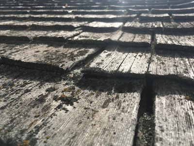 An angled close-up view of weathered wooden shingles, showing a textured surface with visible cracks and patches of moss. The pattern creates a repeating sequence of overlapping rectangles.