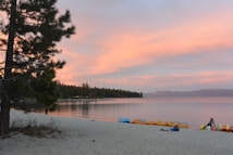 A tranquil lakeside scene at sunset featuring a sandy beach, a line of colorful canoes or kayaks parked along the shore, and calm water stretching toward distant mountains. A pine tree is visible on the left side, and a person lies on the sand, enjoying the peaceful setting. The sky is painted with soft pink and orange hues.