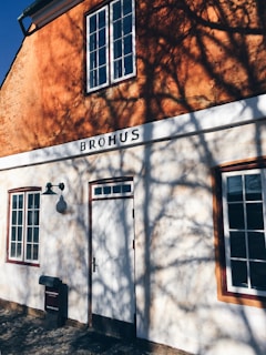 A building with a textured orange wall and white facade casts complex shadow patterns from nearby trees. Large windows are visible, and the word 'BROHUS' is prominently displayed above the door. A vintage lamp and mailbox add to the rustic appearance.