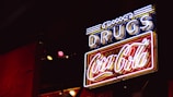 A neon sign with the word 'DRUGS' in blue and a 'Coca-Cola' logo in red, illuminated against a dark background. The sign features a combination of white, blue, and red neon lights, creating a retro or vintage feel.