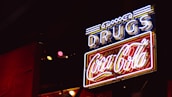 A neon sign with the word 'DRUGS' in blue and a 'Coca-Cola' logo in red, illuminated against a dark background. The sign features a combination of white, blue, and red neon lights, creating a retro or vintage feel.