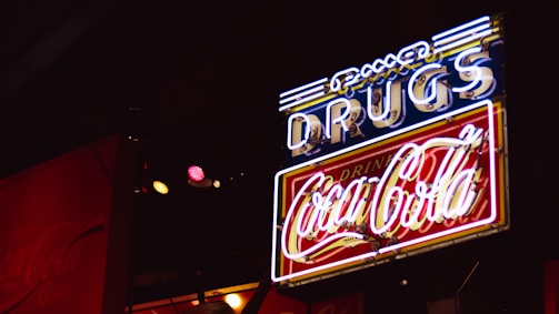 A neon sign with the word 'DRUGS' in blue and a 'Coca-Cola' logo in red, illuminated against a dark background. The sign features a combination of white, blue, and red neon lights, creating a retro or vintage feel.