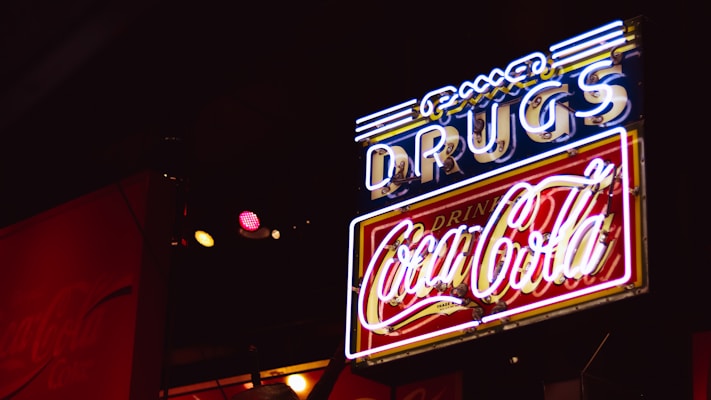 A neon sign with the word 'DRUGS' in blue and a 'Coca-Cola' logo in red, illuminated against a dark background. The sign features a combination of white, blue, and red neon lights, creating a retro or vintage feel.