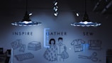 A dimly lit room features two pendant lights with unique designs casting light on a wall. The wall has illustrations and text supporting a creative theme, including words like 'Inspire,' 'Gather,' and 'Sew.' Various drawings depict sewing-related items such as stacked fabric, dress patterns, a sewing machine, scissors, and a shirt with the text 'I made this!' An illustrated couple stands in the middle, holding fabric.