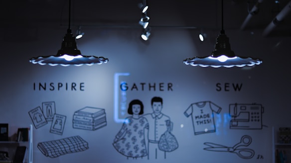 A dimly lit room features two pendant lights with unique designs casting light on a wall. The wall has illustrations and text supporting a creative theme, including words like 'Inspire,' 'Gather,' and 'Sew.' Various drawings depict sewing-related items such as stacked fabric, dress patterns, a sewing machine, scissors, and a shirt with the text 'I made this!' An illustrated couple stands in the middle, holding fabric.