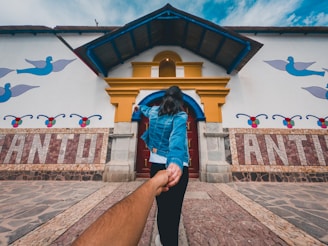 A person in a blue denim jacket is leading another person by the hand towards a colorful building. The building features artistic designs with birds, geometric patterns, and bold letters spelling out 'ANTIOQUIA'. The pathway is made of various stone tiles, and the sky above is blue with some clouds.