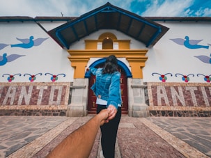 A person in a blue denim jacket is leading another person by the hand towards a colorful building. The building features artistic designs with birds, geometric patterns, and bold letters spelling out 'ANTIOQUIA'. The pathway is made of various stone tiles, and the sky above is blue with some clouds.