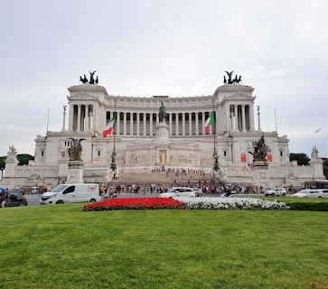 Large, ornate neoclassical building with columns and statues. Two Italian flags are visible at the front. A vibrant garden in the foreground adds color with red and white flowers surrounded by green grass. People are gathered on the steps, and several vehicles are present in front of the structure.