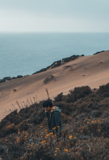 A person in a green jacket stands amidst dense shrubs and blooming yellow flowers on a slope overlooking a vast ocean. The landscape features a sandy hill with patches of vegetation, creating a serene coastal scene under overcast skies.