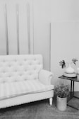 A vintage-style living room setup featuring a tufted couch with striped upholstery. Beside it, a small table holds a white vase with foliage and a candle. A potted plant sits on the floor near the table, and long wooden panels adorn the wall.