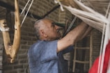 An older man is carefully working with a loom, focused on weaving. He is surrounded by natural materials, including wooden beams and threads. The setting appears rustic and traditional, with brick walls and dim lighting.