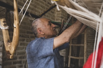 An older man is carefully working with a loom, focused on weaving. He is surrounded by natural materials, including wooden beams and threads. The setting appears rustic and traditional, with brick walls and dim lighting.