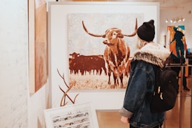 A person wearing a denim jacket with a fur-lined hood and a black beanie is observing a large painting in an art gallery. The painting depicts a longhorn cow in a stylized manner, with abstract floral motifs in the background. Surrounding the main artwork are other paintings and sculptures displayed within the gallery.