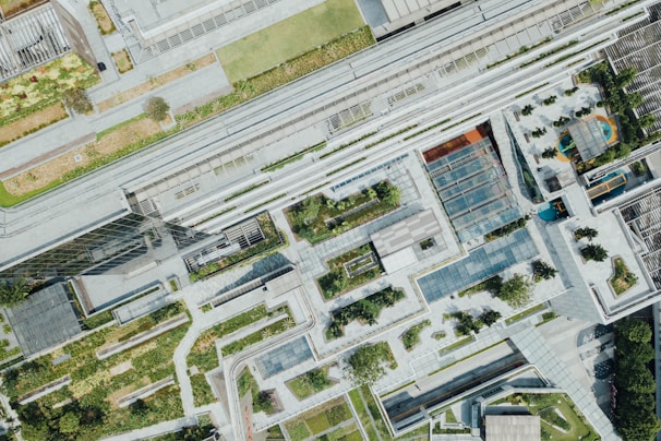 An aerial view of a modern urban landscape featuring a complex arrangement of buildings, green rooftops, and walkways. The area includes numerous garden spaces interspersed between the structures, with pathways and landscaped areas. Glass-covered walkways and solar panels are visible, along with hints of recreational spaces like playgrounds.