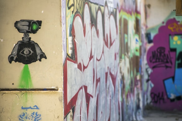 Street art features a stencil of a robot with a single green eye on a weathered wall. The robot has a geometric body and emits a green light downward. Surrounding it, there are various layers of colorful graffiti, including abstract shapes and letters in red, purple, and green tones.