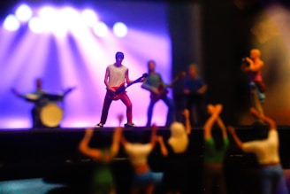 A miniature scene of a rock band performing on stage with a guitarist in focus. The backdrop features bright stage lights casting colorful hues. The foreground shows a small audience with raised hands, emphasizing the lively atmosphere.