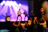 A miniature scene of a rock band performing on stage with a guitarist in focus. The backdrop features bright stage lights casting colorful hues. The foreground shows a small audience with raised hands, emphasizing the lively atmosphere.