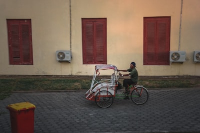 A person rides a traditional tricycle rickshaw beside a beige wall with red shuttered windows. Air conditioning units are mounted on the wall next to the windows. The ground is paved with hexagonal stones, and a red trash bin with a yellow lid is on the side.