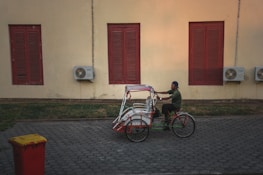 A person rides a traditional tricycle rickshaw beside a beige wall with red shuttered windows. Air conditioning units are mounted on the wall next to the windows. The ground is paved with hexagonal stones, and a red trash bin with a yellow lid is on the side.