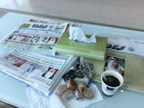 A table holds several newspapers, a pack of tissues, a cup of coffee, keys, and a few packaged cookies. The newspapers feature prominent figures and headlines in Arabic. A tissue box is placed neatly alongside the coffee cup, which is branded. The keys have several keychains attached.