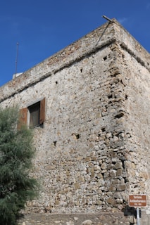 A historical stone tower with a weathered facade, featuring small windows and iron shutters. The structure is set against a clear blue sky, with a small wooden sign indicating 'Torrione Antibarbaresco' and years of construction. A bushy green plant can be seen at the base of the building.