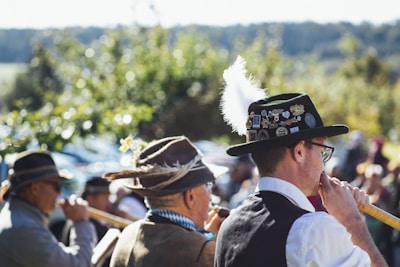 A group of individuals dressed in traditional Bavarian attire, including hats adorned with feathers and pins, are playing long wooden wind instruments at an outdoor gathering. The background features blurred greenery and a crowd of people, suggesting a festive setting.