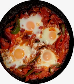 Four eggs are cooked in a skillet with a tomato-based sauce. The dish includes sliced green and red bell peppers, and the eggs have sunny side up yolks surrounded by a red, saucy base.