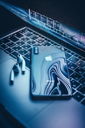 A sleek, modern smartphone with a reflective surface is placed on the keyboard of a laptop. Next to the phone, there are wireless earbuds. The scene is illuminated by a soft blue light, creating a technological and futuristic ambiance.